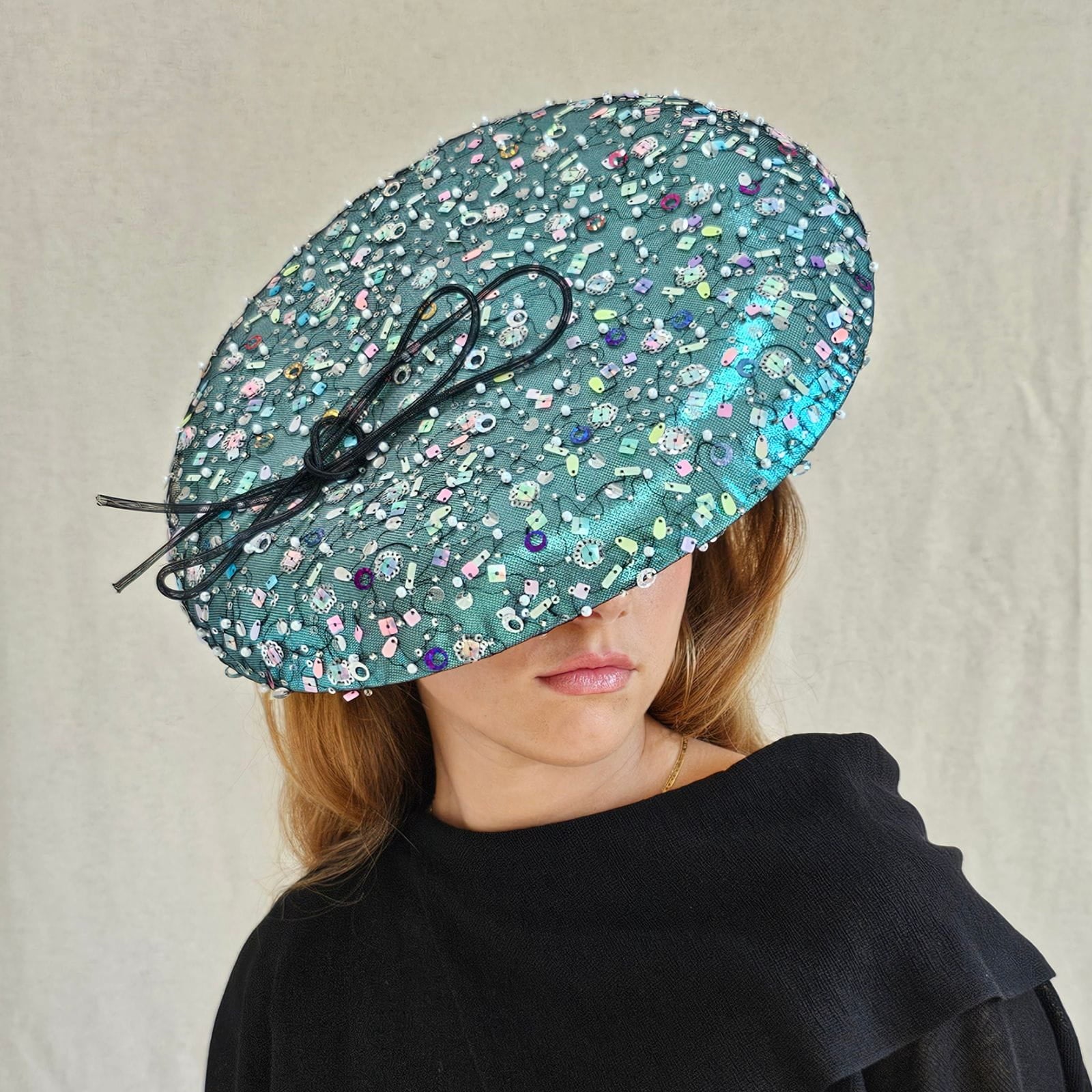 Aqua lamé disc hat with multicolour tulle sequining and elegant black bow, angle view.