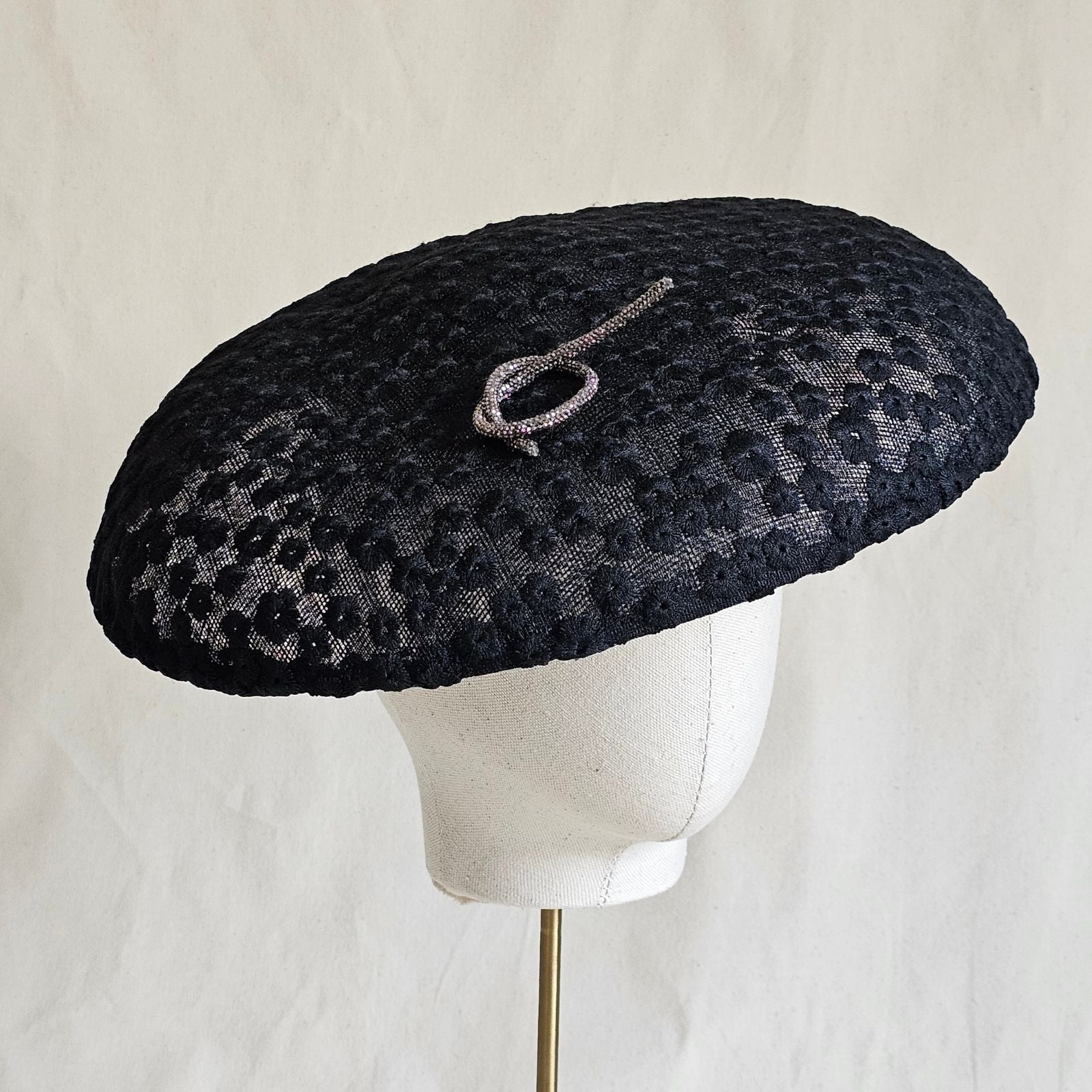 Black disc hat covered in elegant flower lace with silver pretzel knot embellishment, angle view.