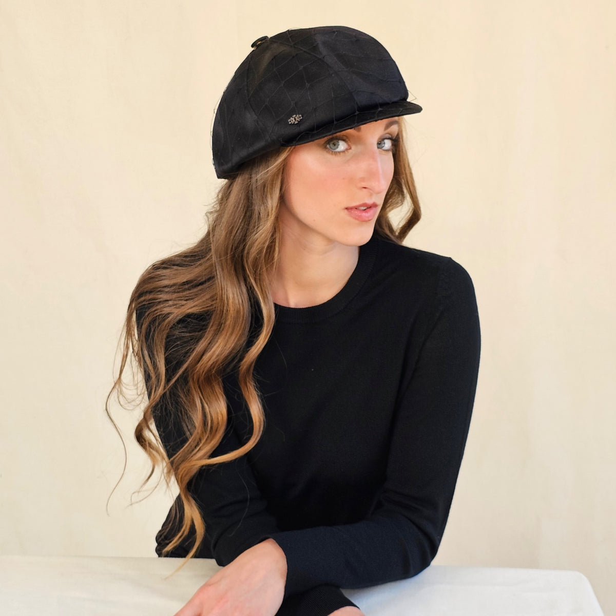 Angled view of black silk satin baker boy cap with veil on a seated
model.