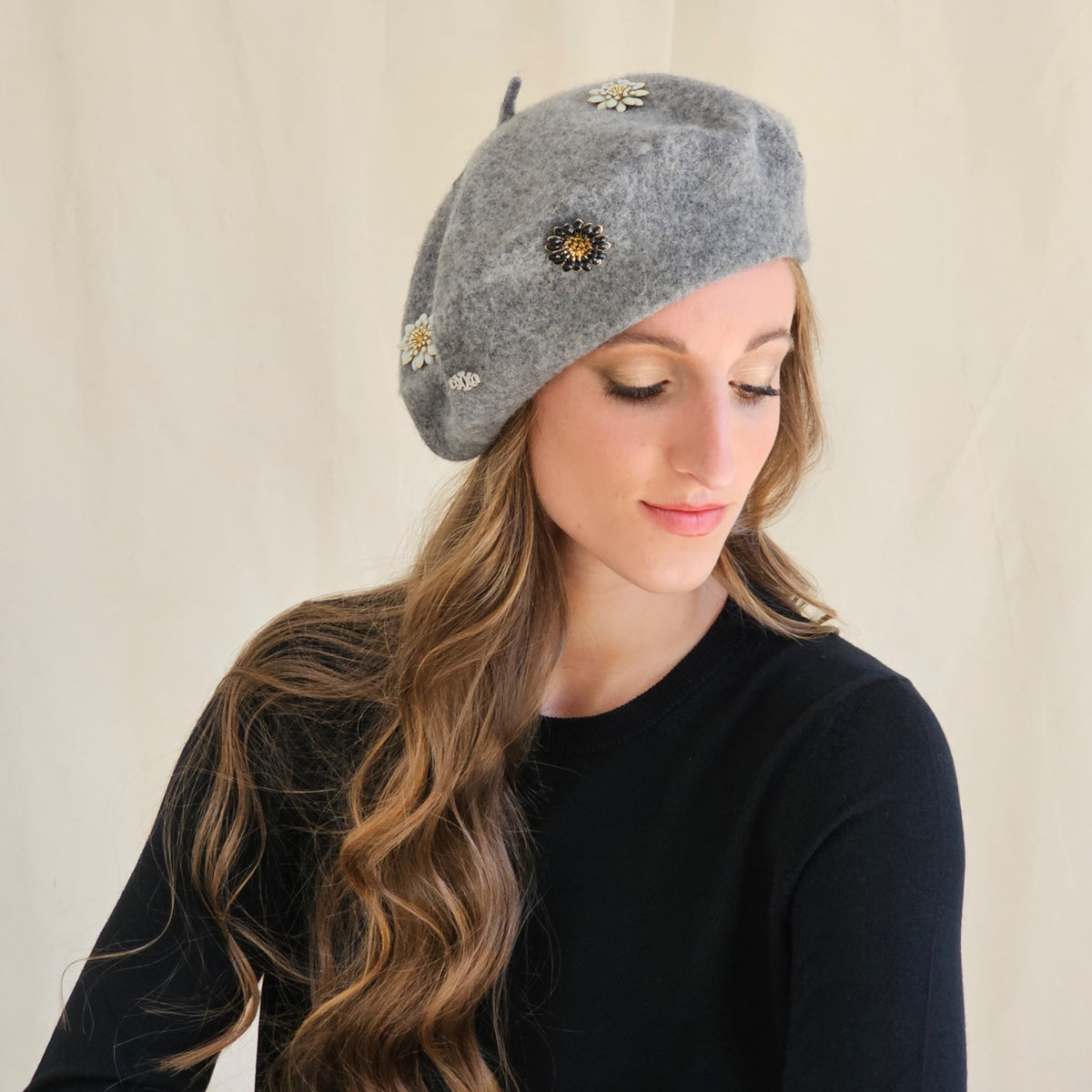 Grey wool beret showing stitched flower and monogram details,
model looking down.